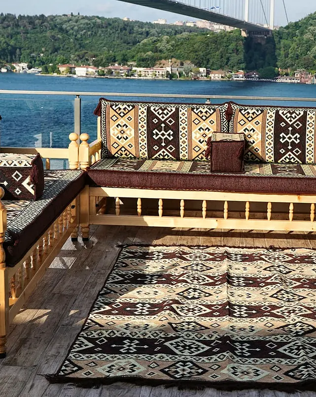 Three pine benches with turned spindles in a U around a kilim rug, with brown and cream kilim-pattern cushions on a waterside terrace
