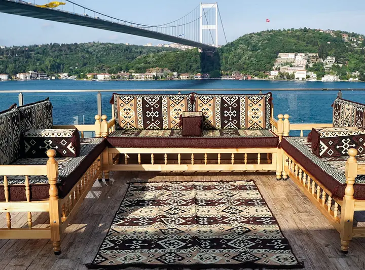 Three pine benches with turned spindles in a U around a kilim rug, with brown and cream kilim-pattern cushions on a waterside terrace