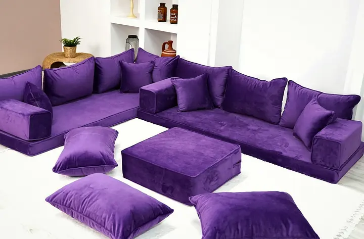 Purple velvet L-shaped floor sofa with a square ottoman and floor cushions, set into the corner of a white room