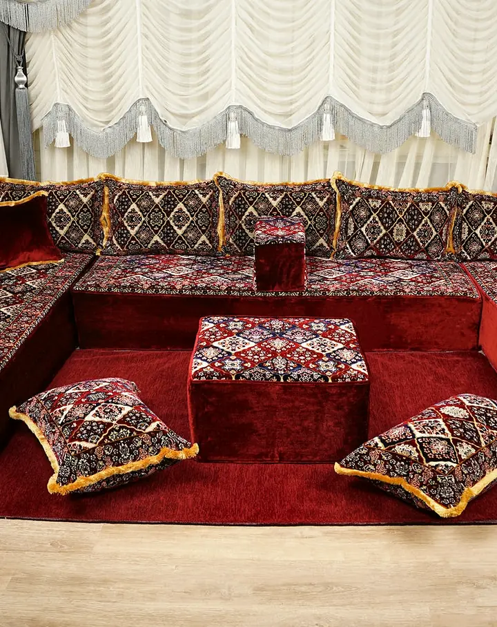 Red seating surrounds a square ottoman on a red rug, with floral diamond backrests, gold fringes and cream gathered curtains