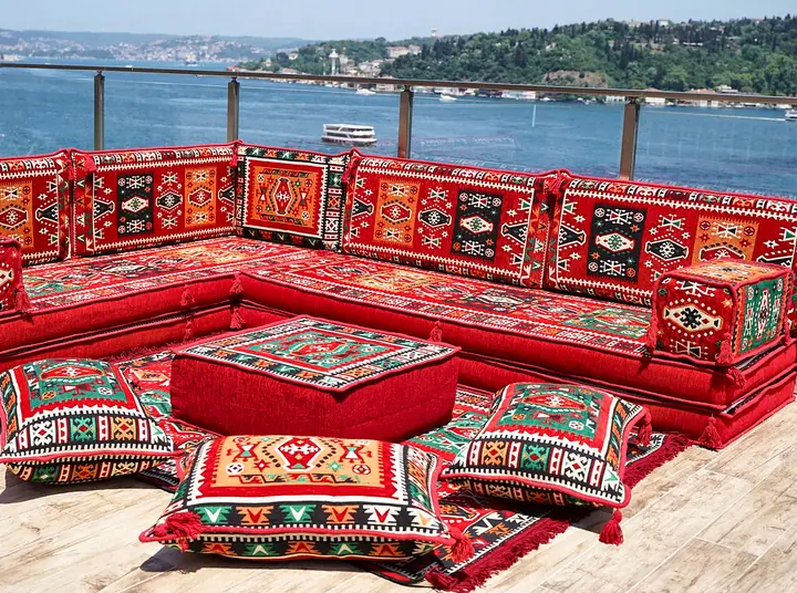 Red kilim-pattern floor sofa wrapping a terrace corner, with a square ottoman and floor pillows and the sea behind