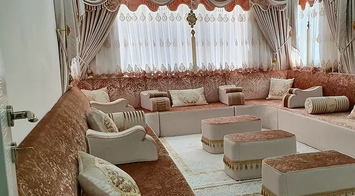 Rose-pink damask majlis along three walls with cream bases, rectangular ottomans edged in gold tassels, a white rug and swag curtains