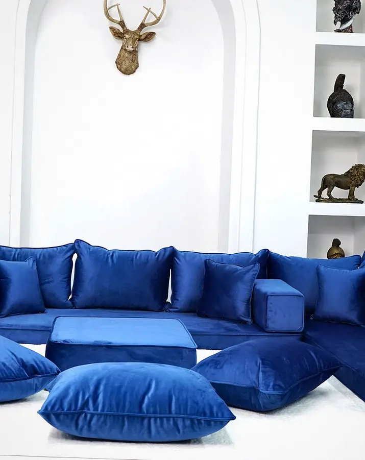 Royal blue velvet floor sofa on three sides of a white room, with an ottoman, floor pillows and an arched niche with shelving
