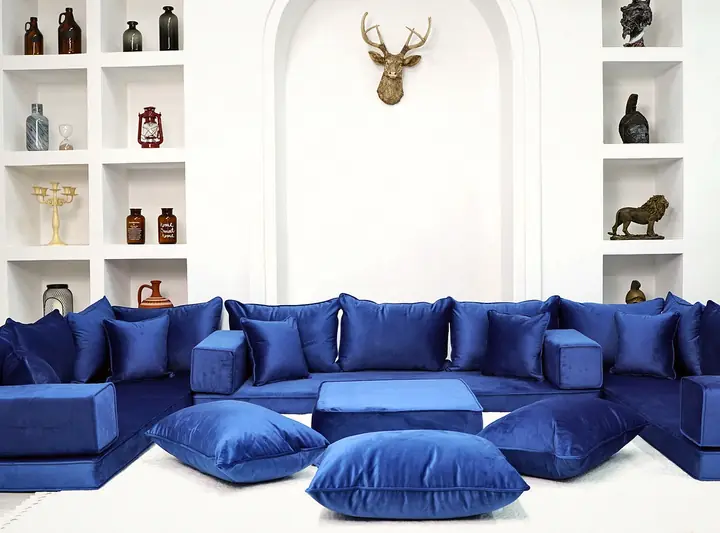 Royal blue velvet floor sofa on three sides of a white room, with an ottoman, floor pillows and an arched niche with shelving