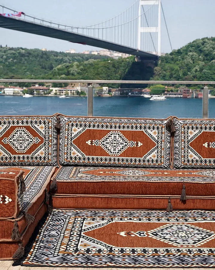 Rust brown L-shaped floor sofa set with kilim-diamond backrests and a matching rug on a glass-railed terrace by the water