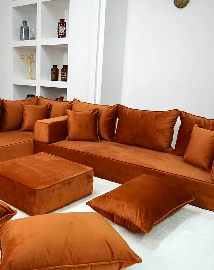 Rust orange velvet L-shaped floor majlis with matching backrests, a square ottoman and three floor pillows on a white rug