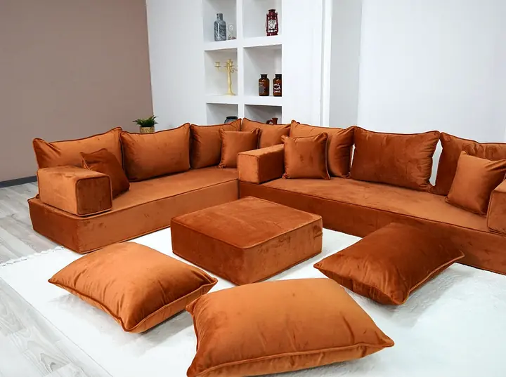 Rust orange velvet L-shaped floor majlis with matching backrests, a square ottoman and three floor pillows on a white rug