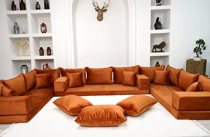 Rust orange velvet U-shaped floor sofa with square backrests, bolster armrests and three floor pillows on a white fringed rug