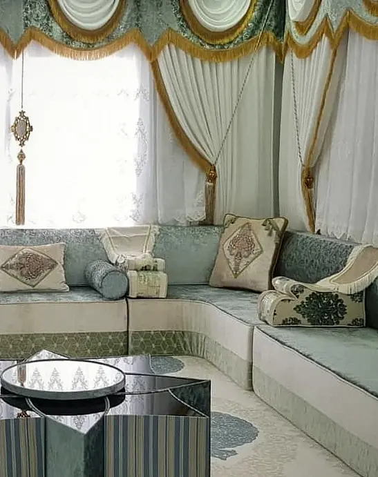 Sage green jacquard majlis along three walls under tall windows with gold-fringed valances, a striped star-shaped table and a cream rug