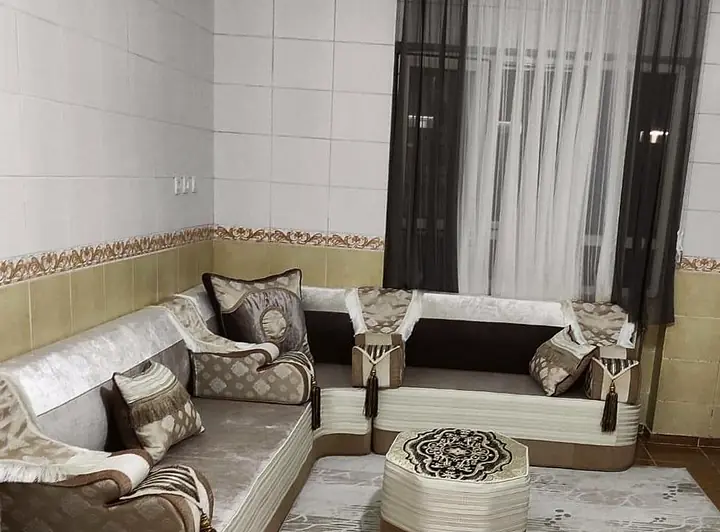 Silver crushed-velvet majlis turning a corner between a tiled wall and a curtained window, with a cut-corner pouf on a pale patterned rug
