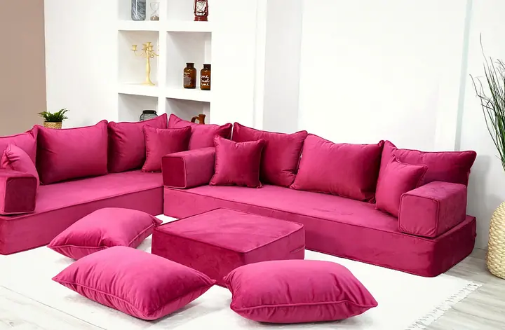 Pink velvet corner floor couch with square backrests and throw pillows, a pink ottoman and floor cushions on a white rug