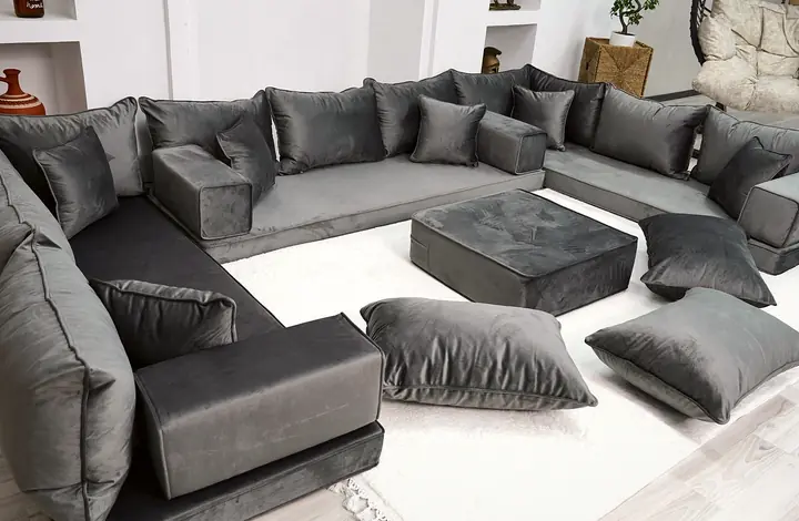 Gray velvet floor sectional on three walls with darker seat bases, an ottoman and floor pillows, viewed along one side