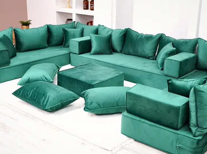 Angled view of an emerald green velvet U set with block armrests, a green ottoman and floor pillows across the floor