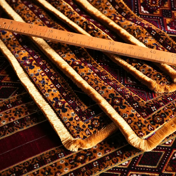 Stacked panels of brown and maroon patterned upholstery fabric with gold fringe edges, a wooden ruler laid across them