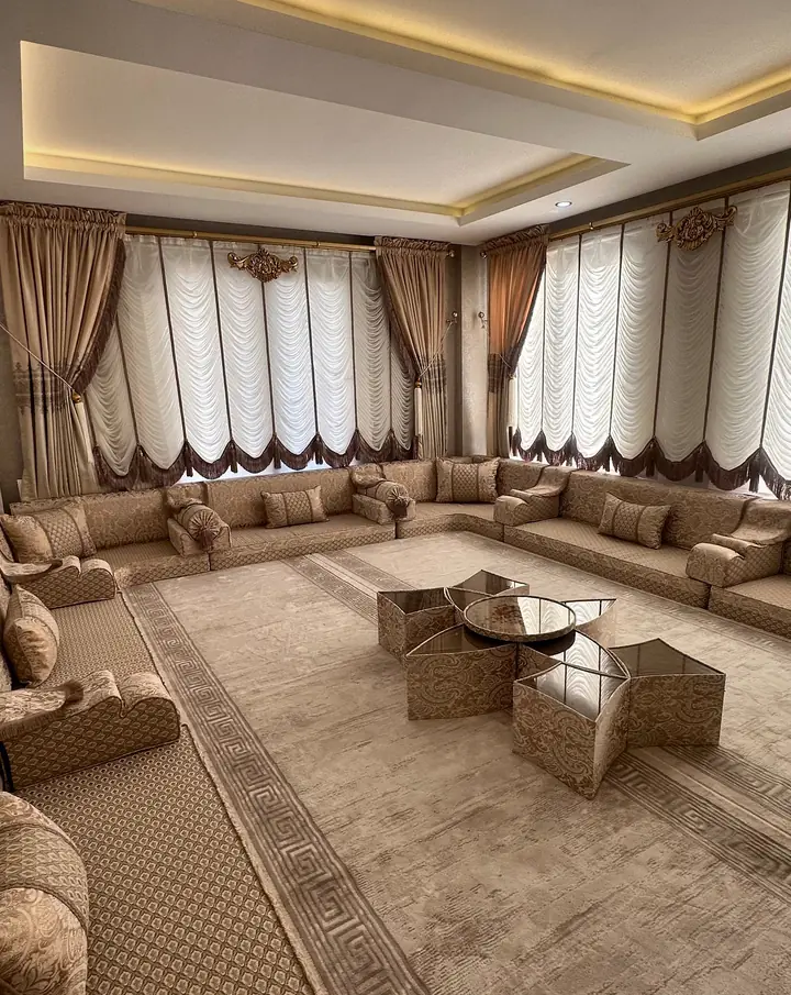 Tan velvet majlis seating along three walls of a long room with round mirrored tables, a matching carpet and cove ceiling lights