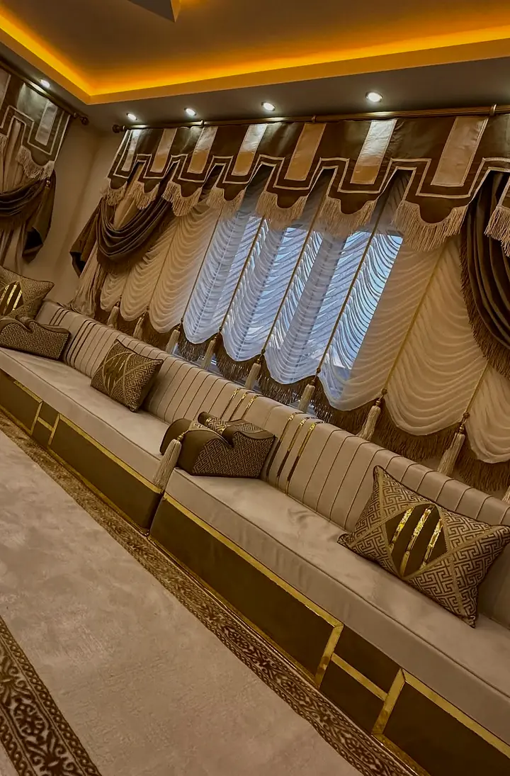 Low view along cream channel-stitched backrests and a mocha base framed in gold lines, with a Greek-key pillow and fringed valance