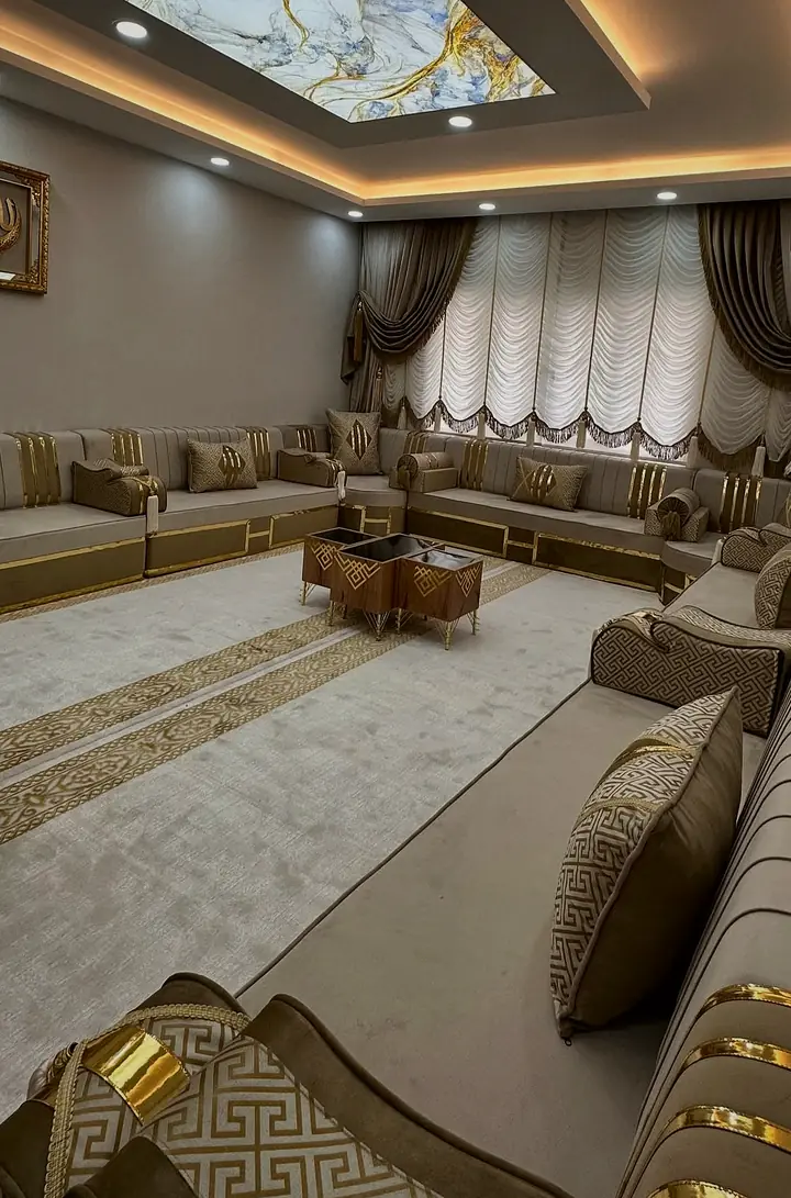 Corner where a Greek-key bolster with a gold band meets taupe backrests crossed by polished gold bars, beside a pale rug