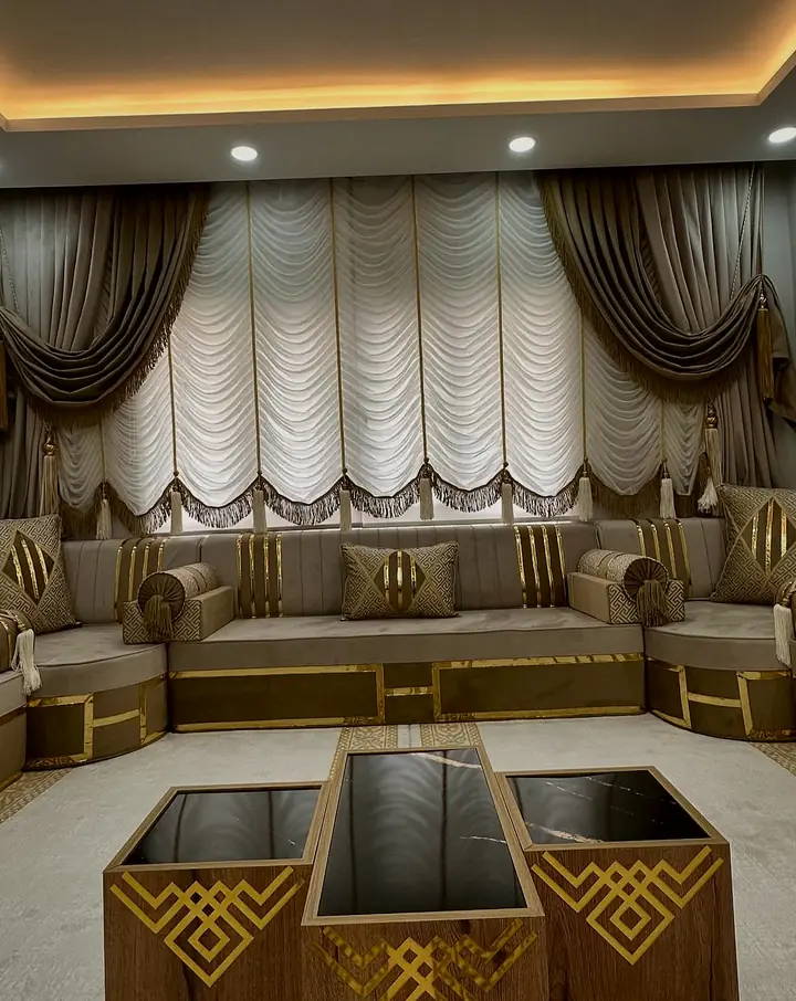 Taupe majlis on three walls with gold-banded backrests and Greek-key pillows below white festoon blinds and dark swag curtains
