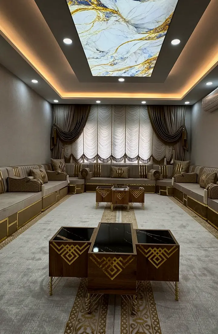 Long view of a taupe and gold majlis room under a marble-print lit ceiling panel, with nesting tables on a Greek-key runner rug