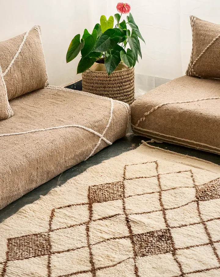 Two taupe Moroccan-style floor couches meeting in a corner, with white cord lines, lumbar pillows and a cream diamond-pattern rug