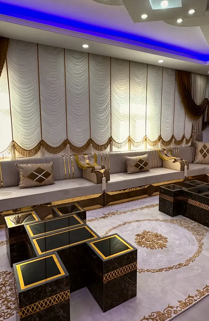 Taupe majlis under white balloon curtains with brown swags, gold armrests and two sets of dark nesting tables on a medallion rug