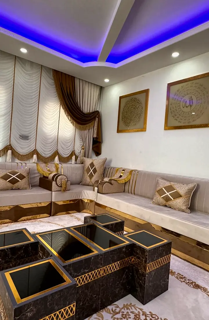 Corner of a taupe velvet majlis with diamond-pattern pillows and gold-trimmed bases, beside nesting tables with black glass tops