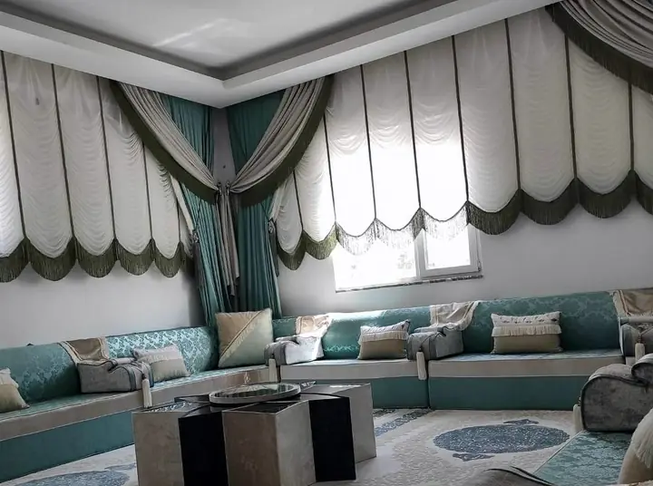 Teal and sage majlis along the walls of a large room, with scalloped Austrian blinds, a star-shaped table and a patterned rug