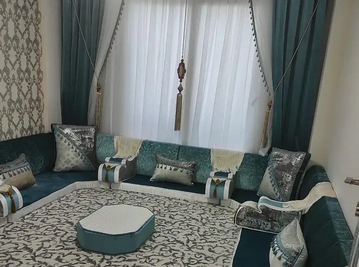Deep teal and silver crushed-velvet majlis around three walls, with an octagonal ottoman on a cream scroll-pattern rug and teal curtains