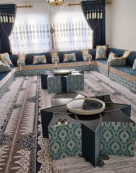 Wide view of a U-shaped teal velvet majlis with cream edging and jacquard bands, two star-shaped tables, a patterned rug and navy curtains