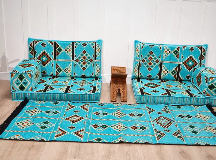 Two turquoise kilim floor seats with a matching flat-woven rug and a carved wooden side table against a white paneled wall