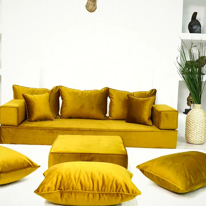 Mustard velvet floor sofa with five back pillows, a matching ottoman and three floor pillows on a white rug in a white room
