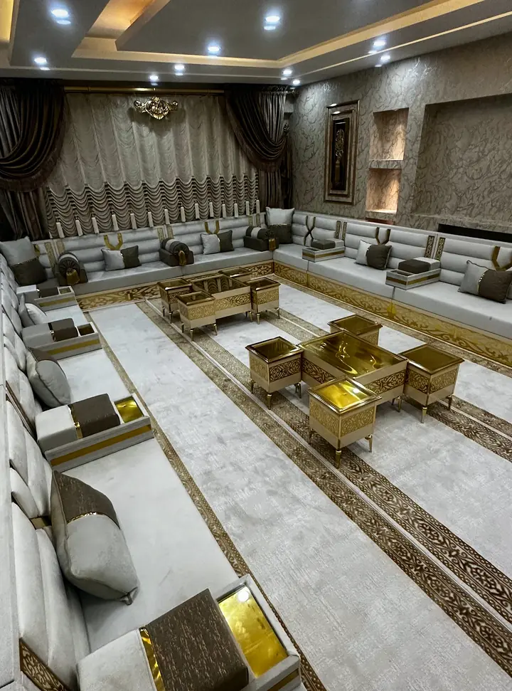 White and gray majlis lining a long room, with gold armrest panels, taupe armrest pads and gold-framed tables on a cream rug
