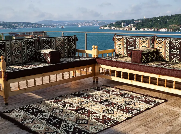 Two light wooden benches in an L with turned spindles, dark brown seat cushions, kilim-pattern backrests and a flat-woven rug on a terrace