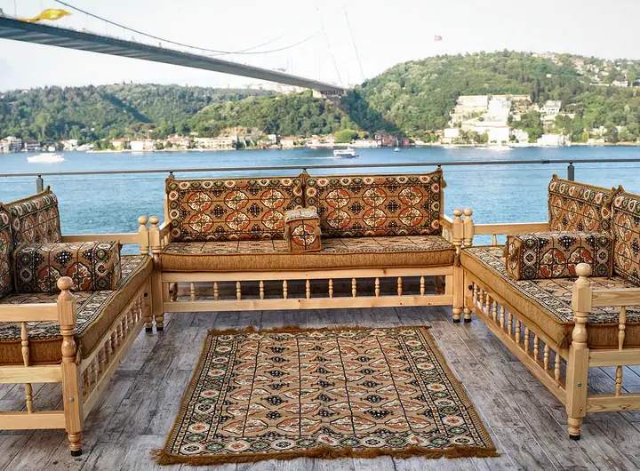 U-shaped wooden bench majlis with gold Afghan-pattern cushions and a matching rug, set outdoors below a suspension bridge